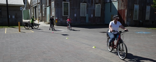 Woman rides around low plastic cones on asphalt surface with three riders in the background in front of older wooden buildings.