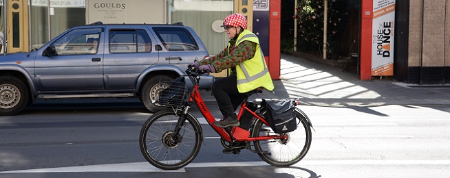 Older woman wearing red and white dotty helmet and yellow high-vis vest rides on road on a red strep through e-bike.