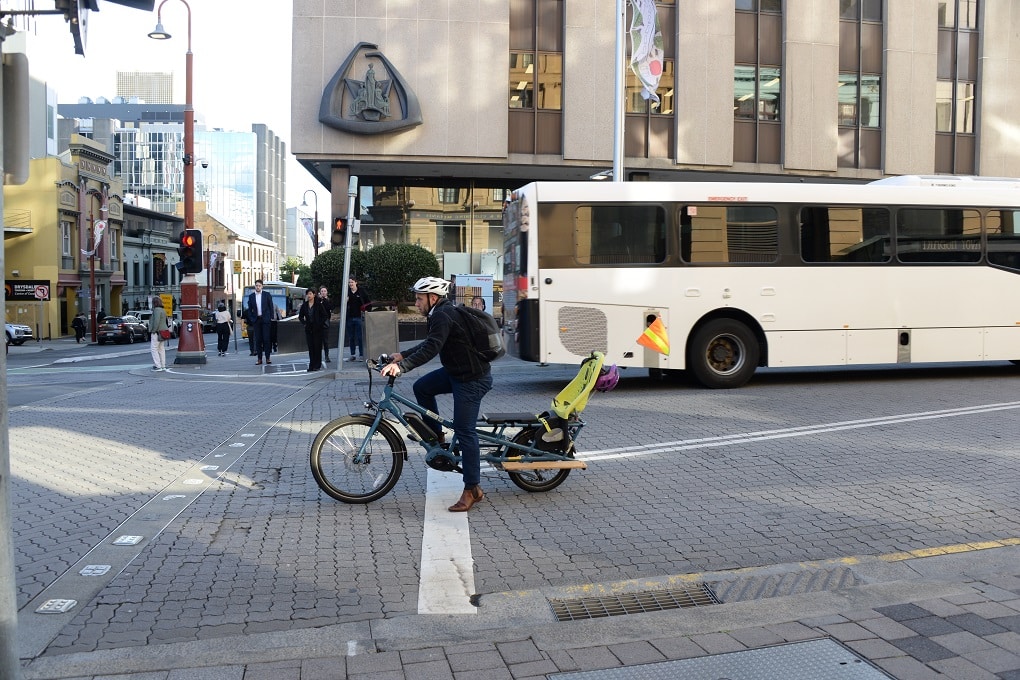A man riding a cargo bike with a child seat on the bike waits at the lights on Elizabeth at Collins Street.