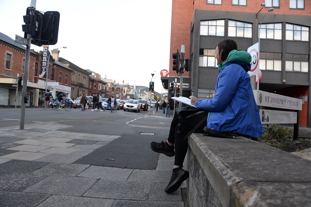 A woman wearing a long blue raincoat with hood sits cross legged on a stone fence looking towards the intersection of Macquarie and Harrington streets.