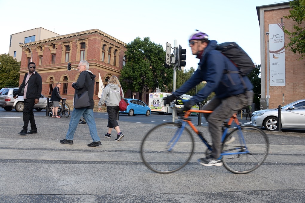 A man riding an orange bike is slightly blurred in the foreground as he passes people walking towards the Argyle and Davey streets intersection.