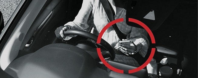 Black and white photo of person driving with a mobile phone in one hand and red circle around the hand with the phone.