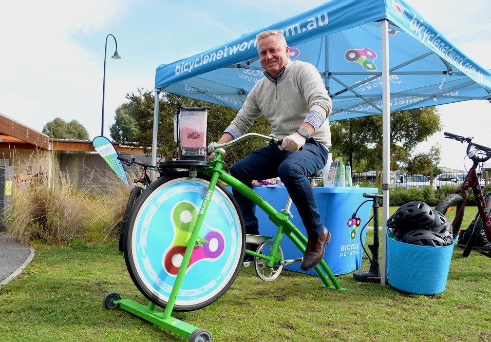 Man wearing natural wool jumper and jeans sits on a green bicycle with a food blender above the front wheel which has a Bicycle Network logo taking up the wheel space.