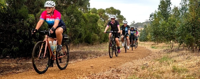 A group of riders in single file ride along a gravel track with bush to their right.