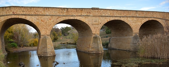 A sandstone bridge with an 1864 date in the middle crosses a river with arched spans and people in the background walking on paths.