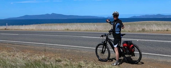 Man on a bicycle stopped by the side of a road along the coast with blue sky and blue sea in the background.