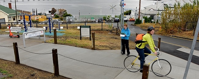 A woman wearing a blue Bicycle Network tshirt stands next to a concrete path with clipboard as a woman weariting a bright yellow raincoat rides past, the background shows industrial maritime buildings.